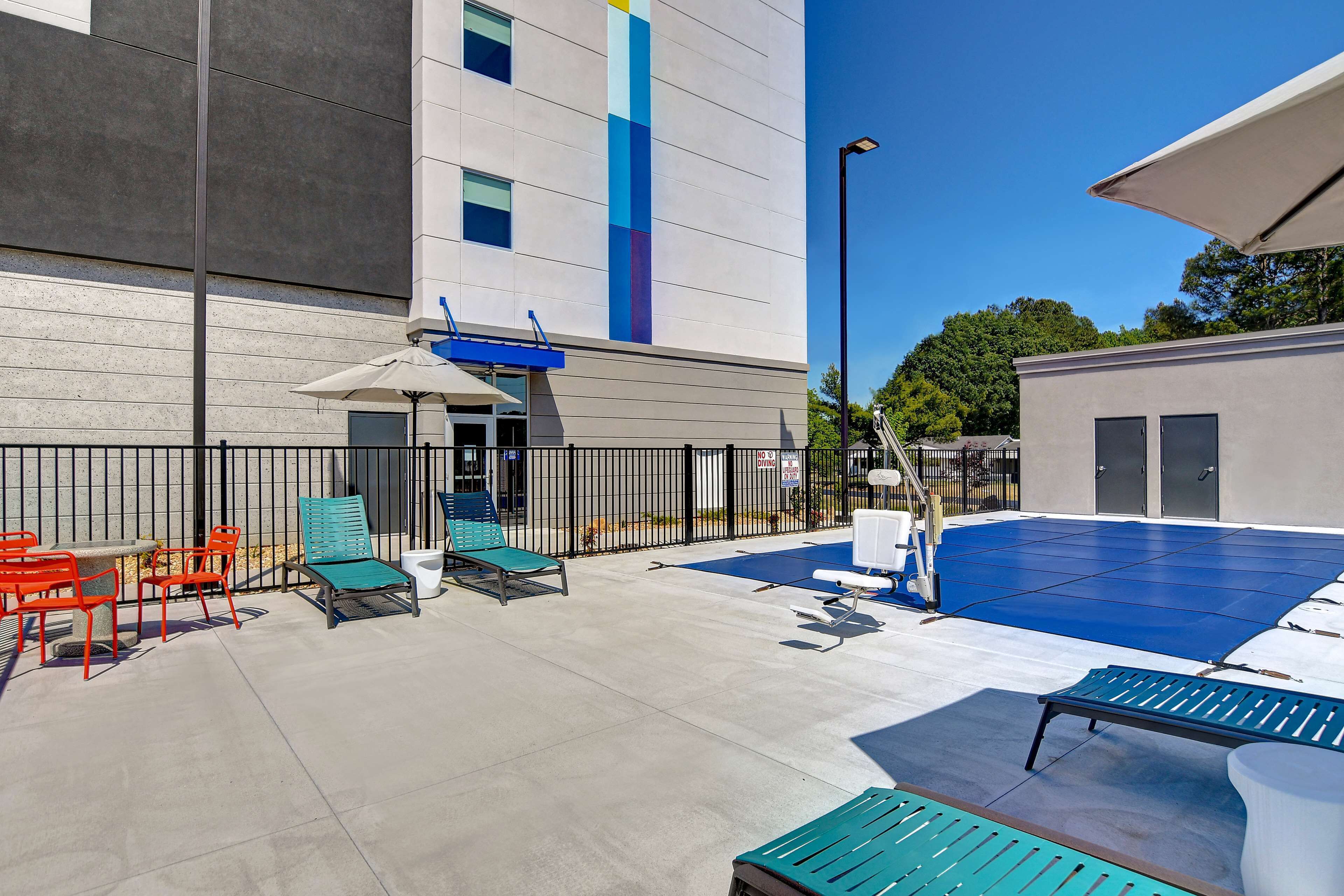 An outdoor pool area with lounge chairs and a table, surrounded by a fence and a building with a blue stripe.