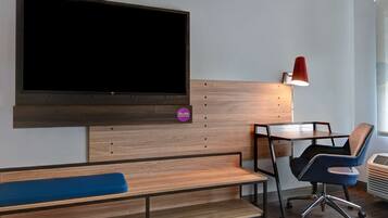 A modern room with a large flat-screen TV mounted on the wall, a desk with a chair, a bench, and a desk lamp.
