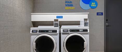 A laundry area with two front-loading washing machines, a water heater sign, and a wall with a laundry instruction sign.