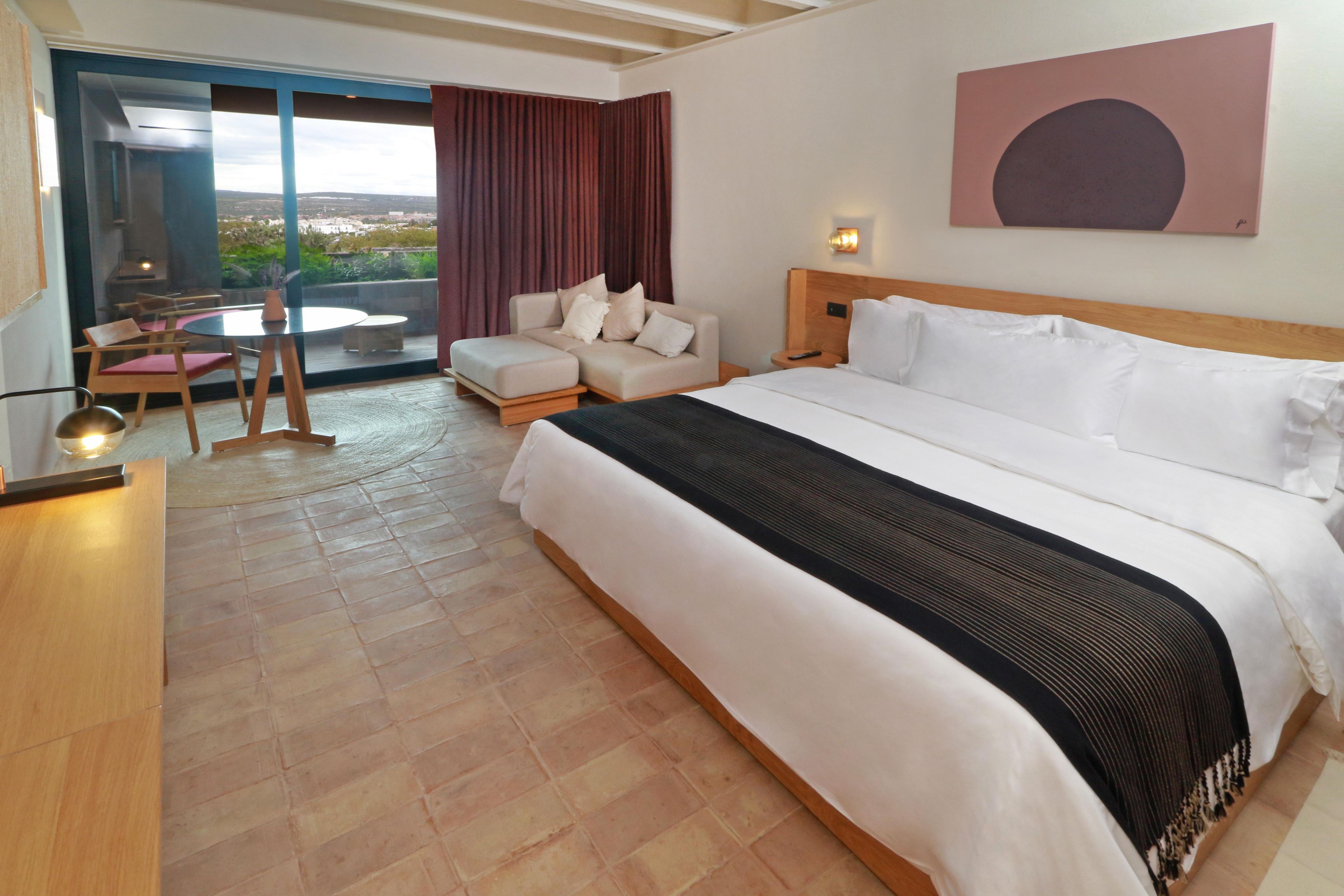 A hotel room with a large bed, a sofa, a dining table, and a view of the outdoors.