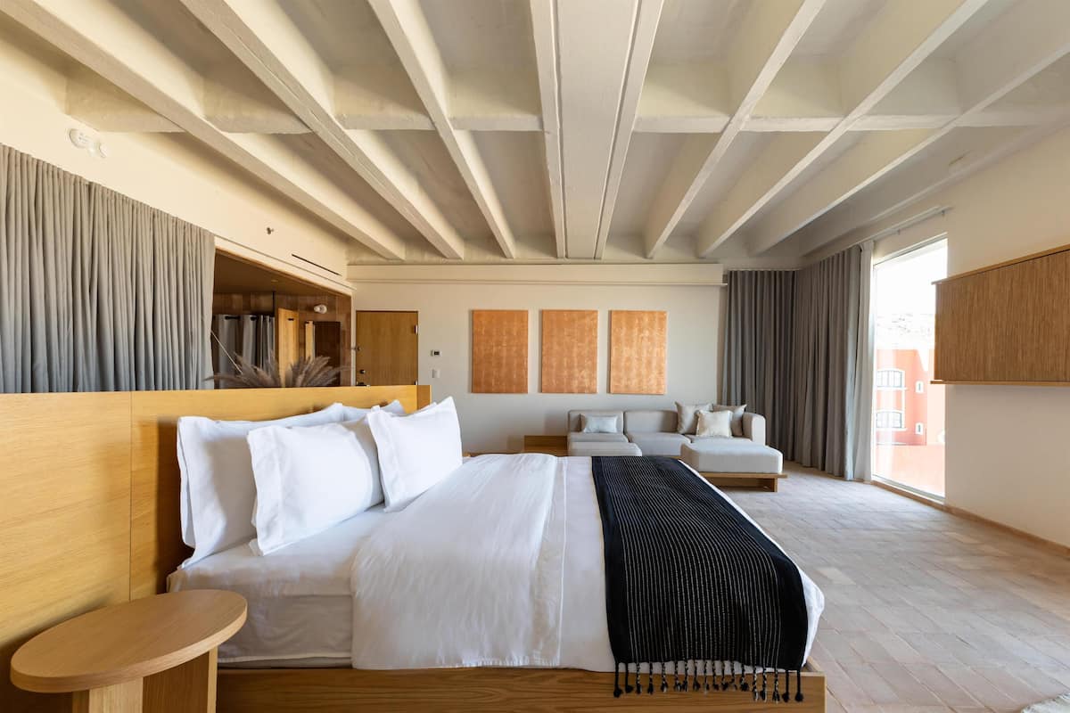 A modern hotel room with a large bed, a wooden headboard, a small wooden table, and a view of the city through large windows.