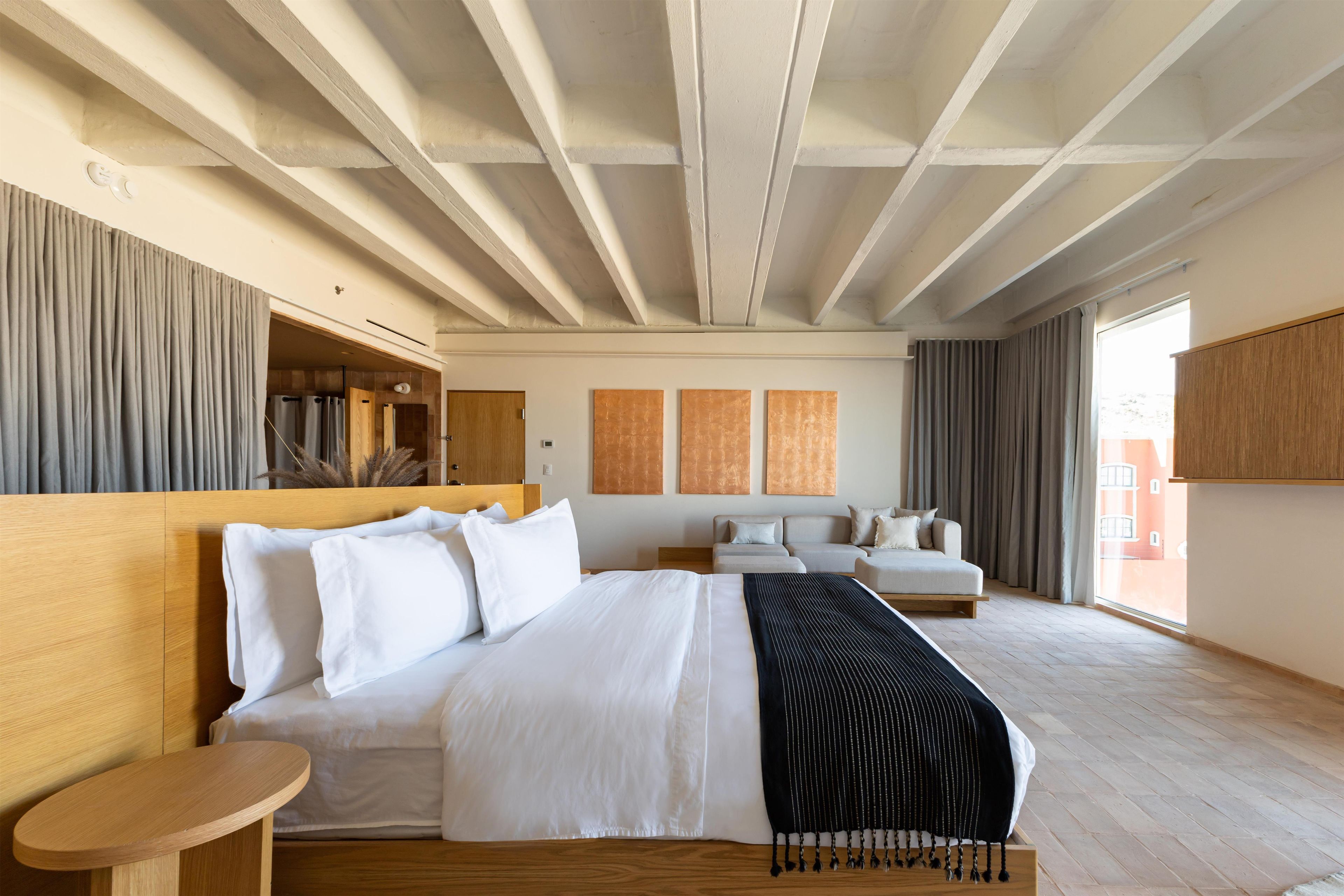 Une chambre d’hôtel avec un grand lit, une tête de lit en bois, une table de chevet, un canapé et une grande fenêtre.