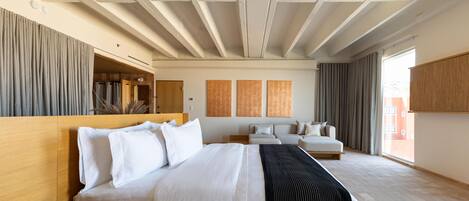 Una habitación de hotel con una cama grande, cabecera de madera, mesita de noche, sofá y un ventanal amplio.