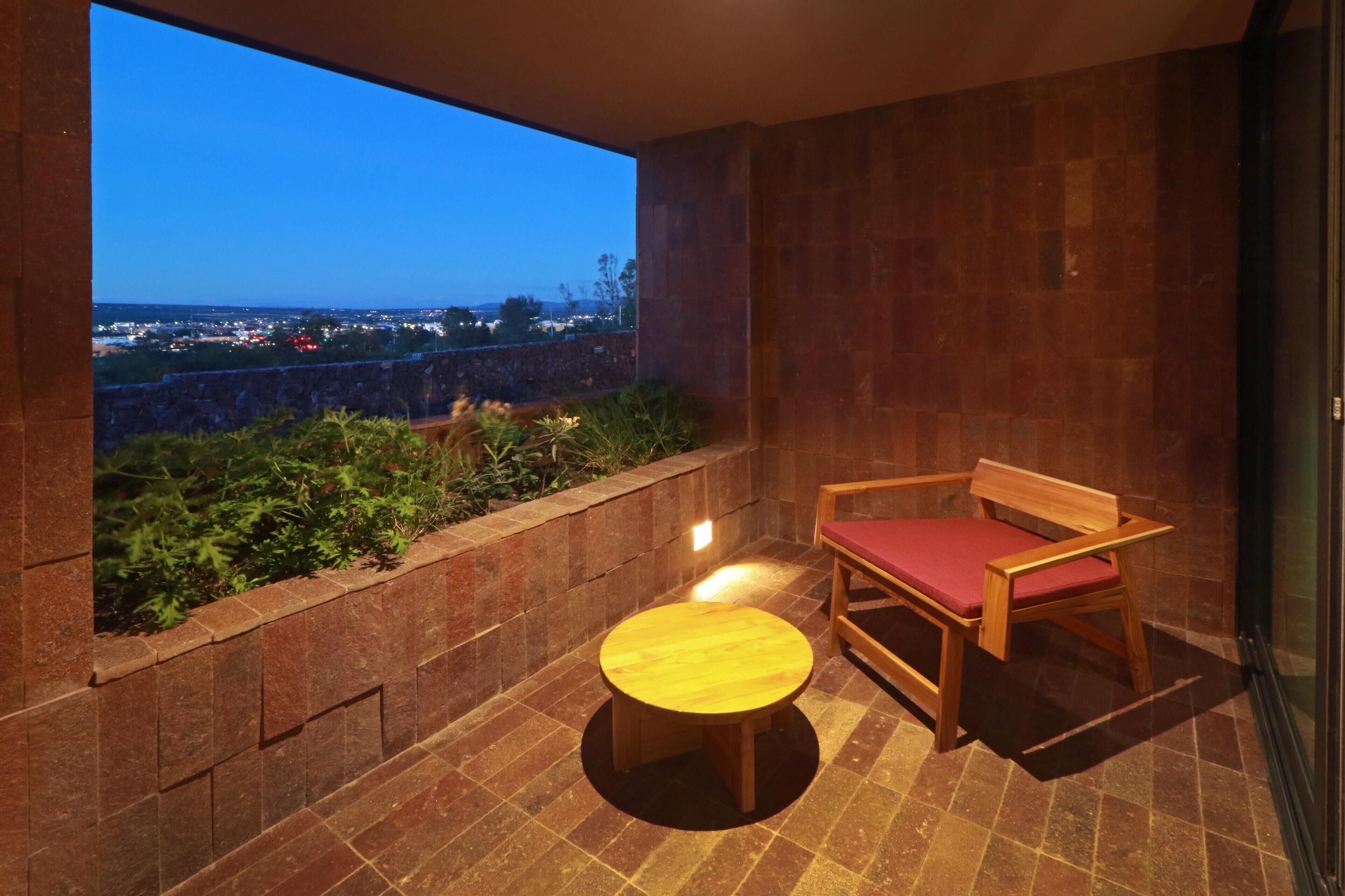 A balcony with a view of a cityscape at dusk, a wooden chair with a red cushion, and a small round table.