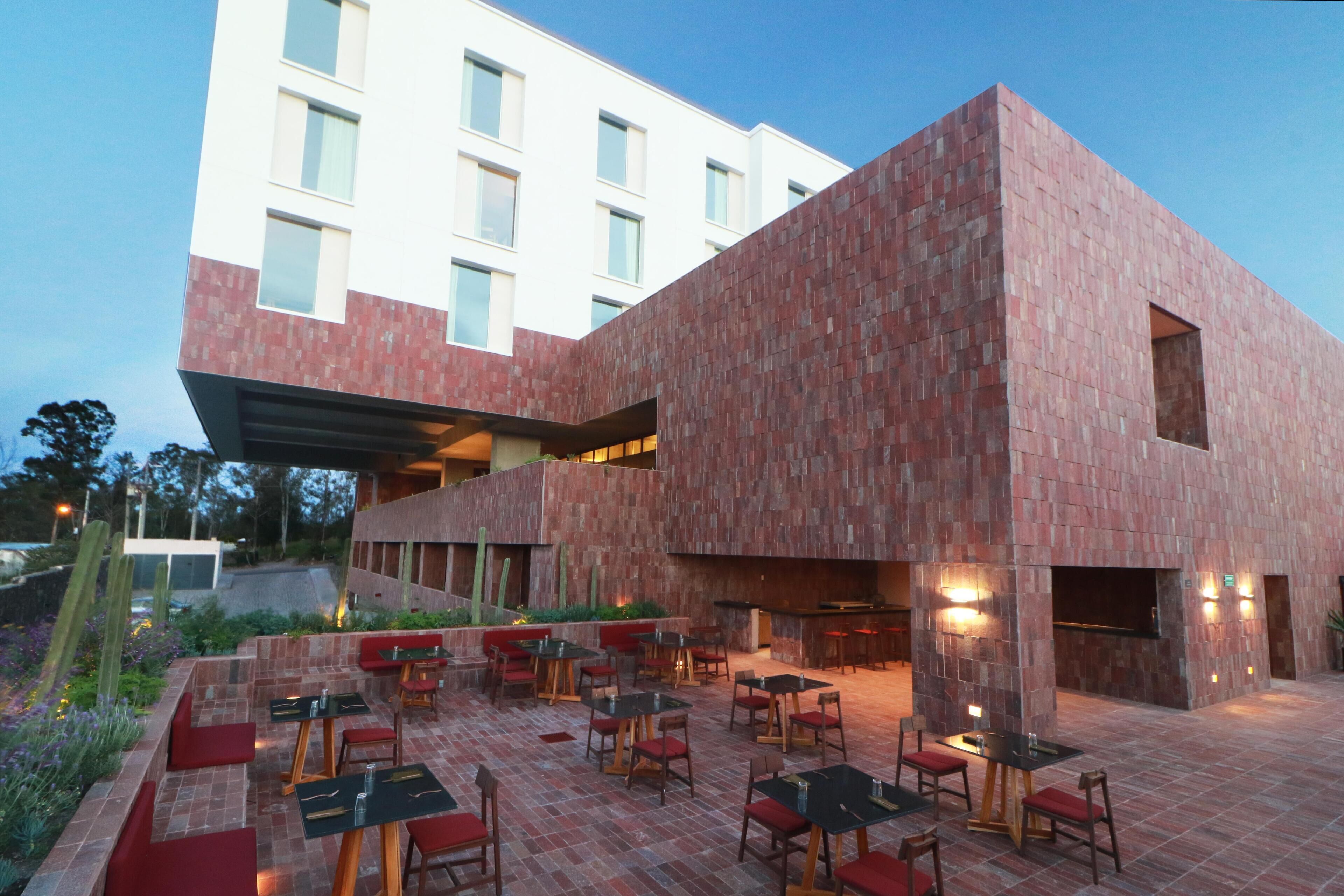 Un bâtiment moderne à la façade en briques rouges, doté d’un espace extérieur aménagé avec des tables et des chaises, ainsi que d’un bar.