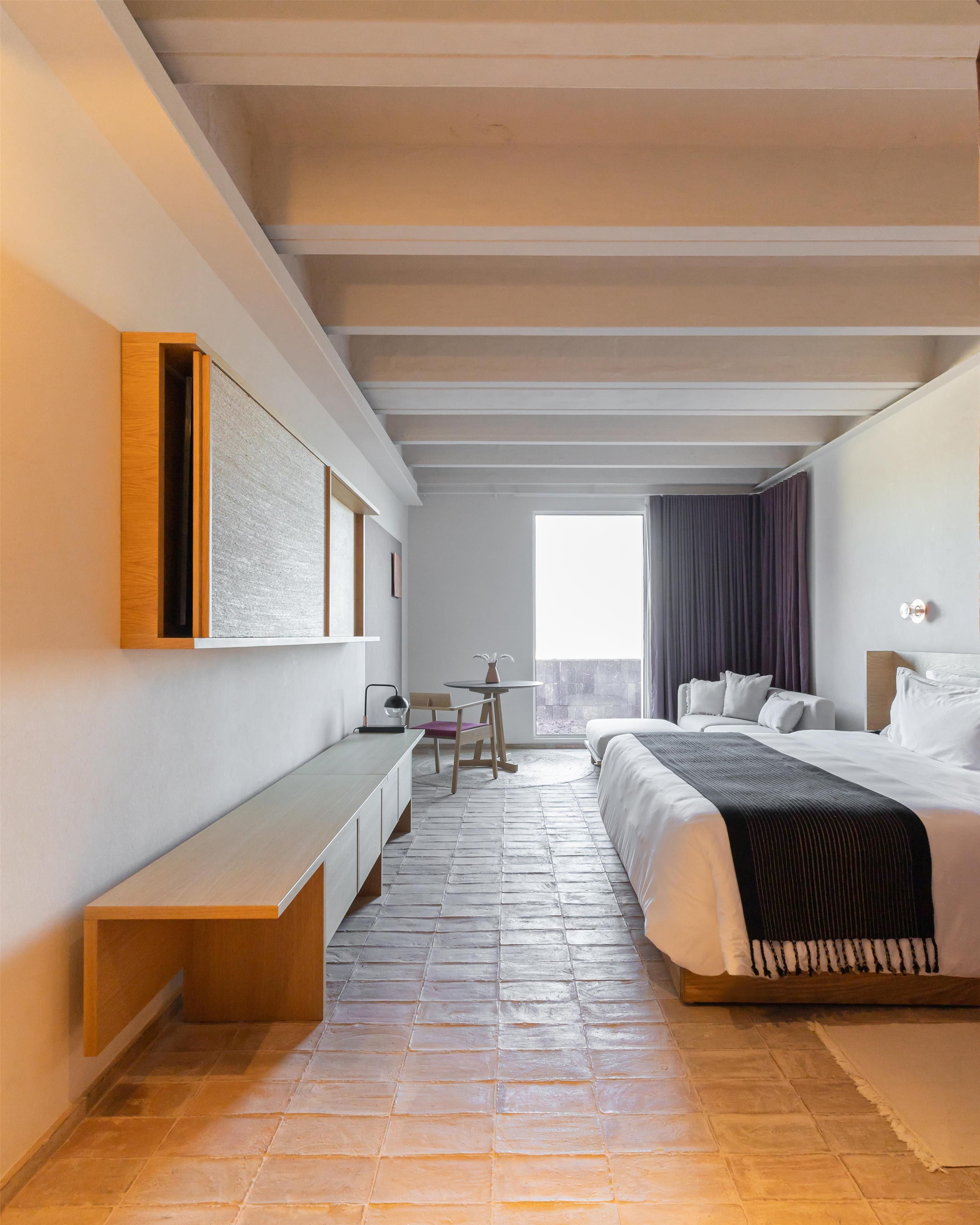 A modern hotel room with a bed, bedside table, bench, and a view of the outdoors.