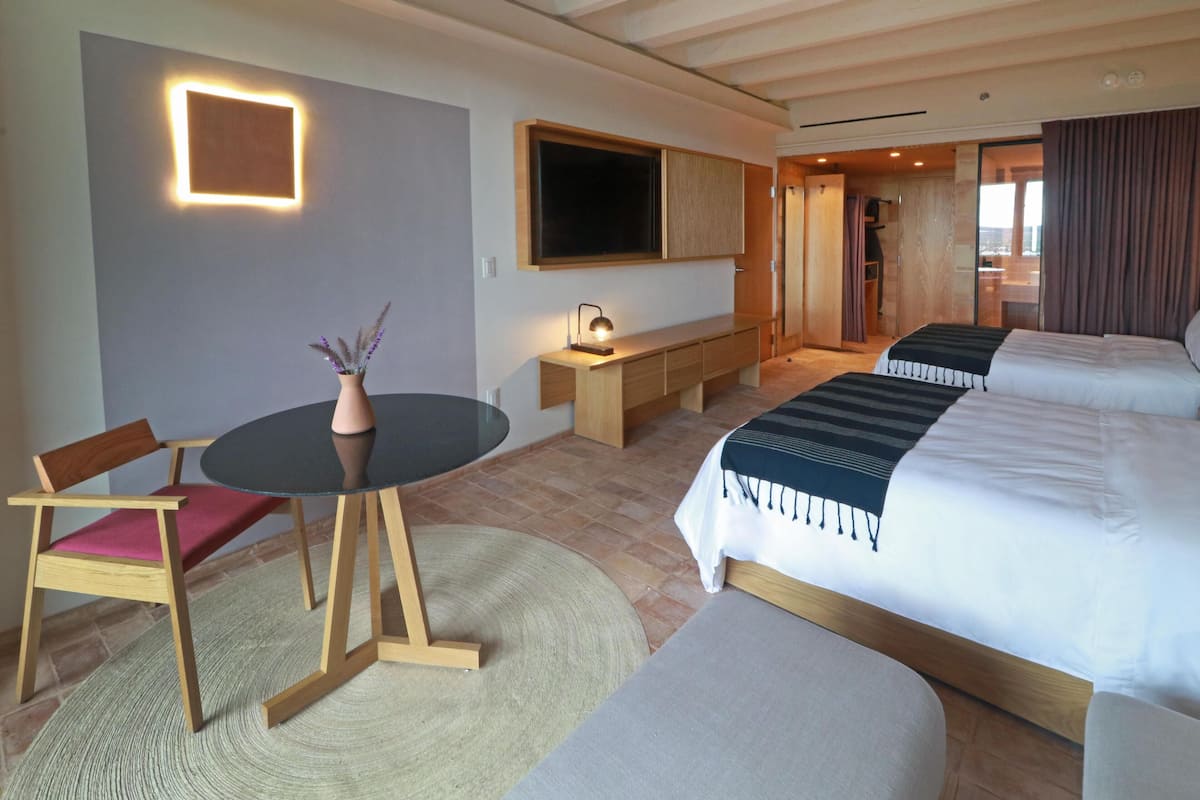 A hotel room with a large bed, a desk with a lamp, a chair, a round rug, and a TV mounted on the wall.