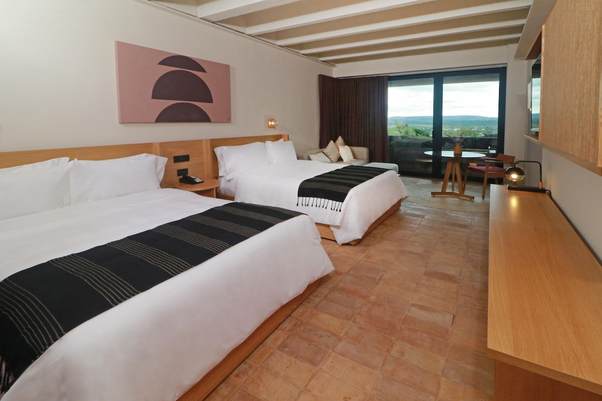 Een hotelkamer met twee bedden, een bureau, een stoel, een raam met uitzicht en een schilderij aan de muur.