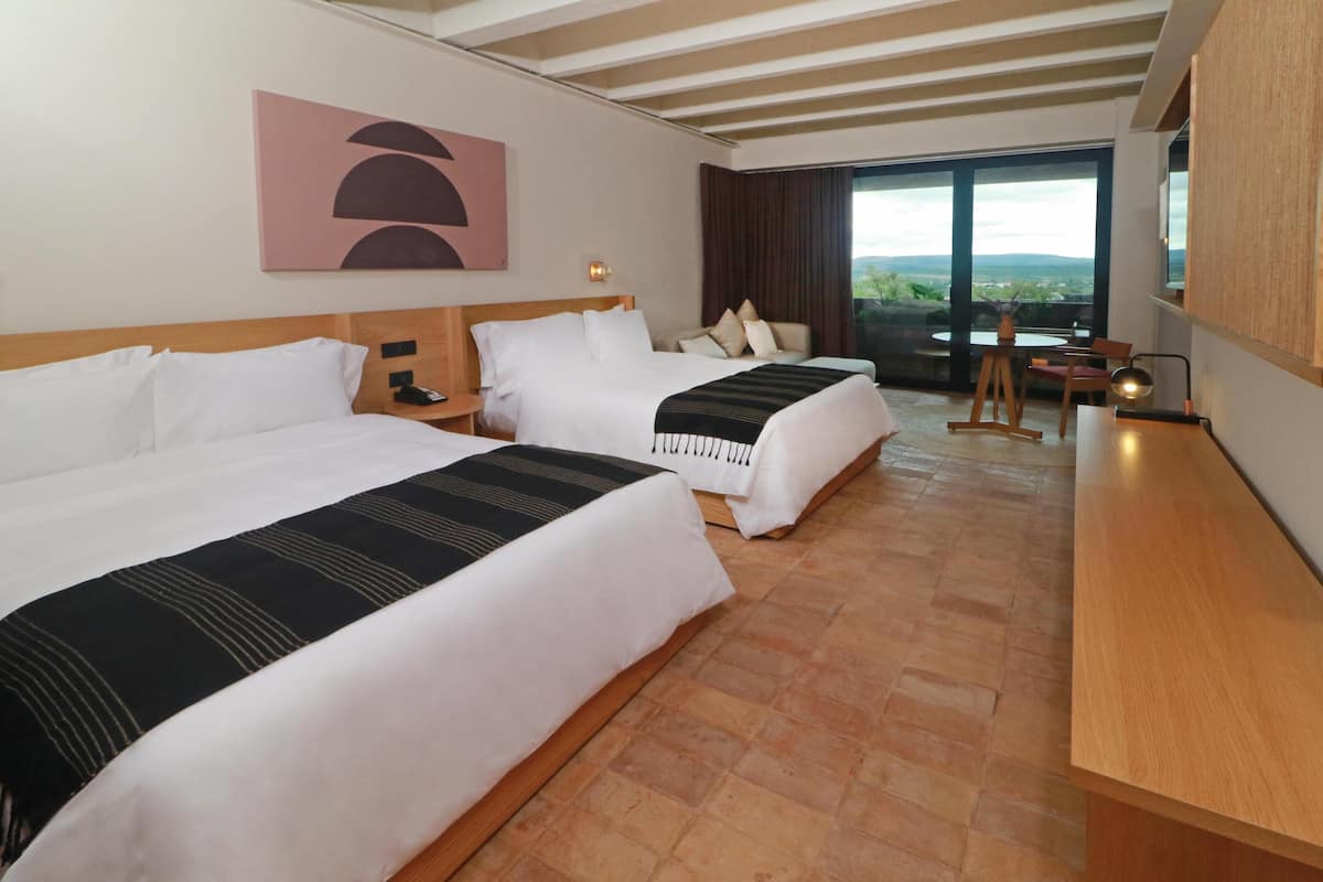 A hotel room with two beds, a wooden desk, a painting on the wall, and a view through the window.