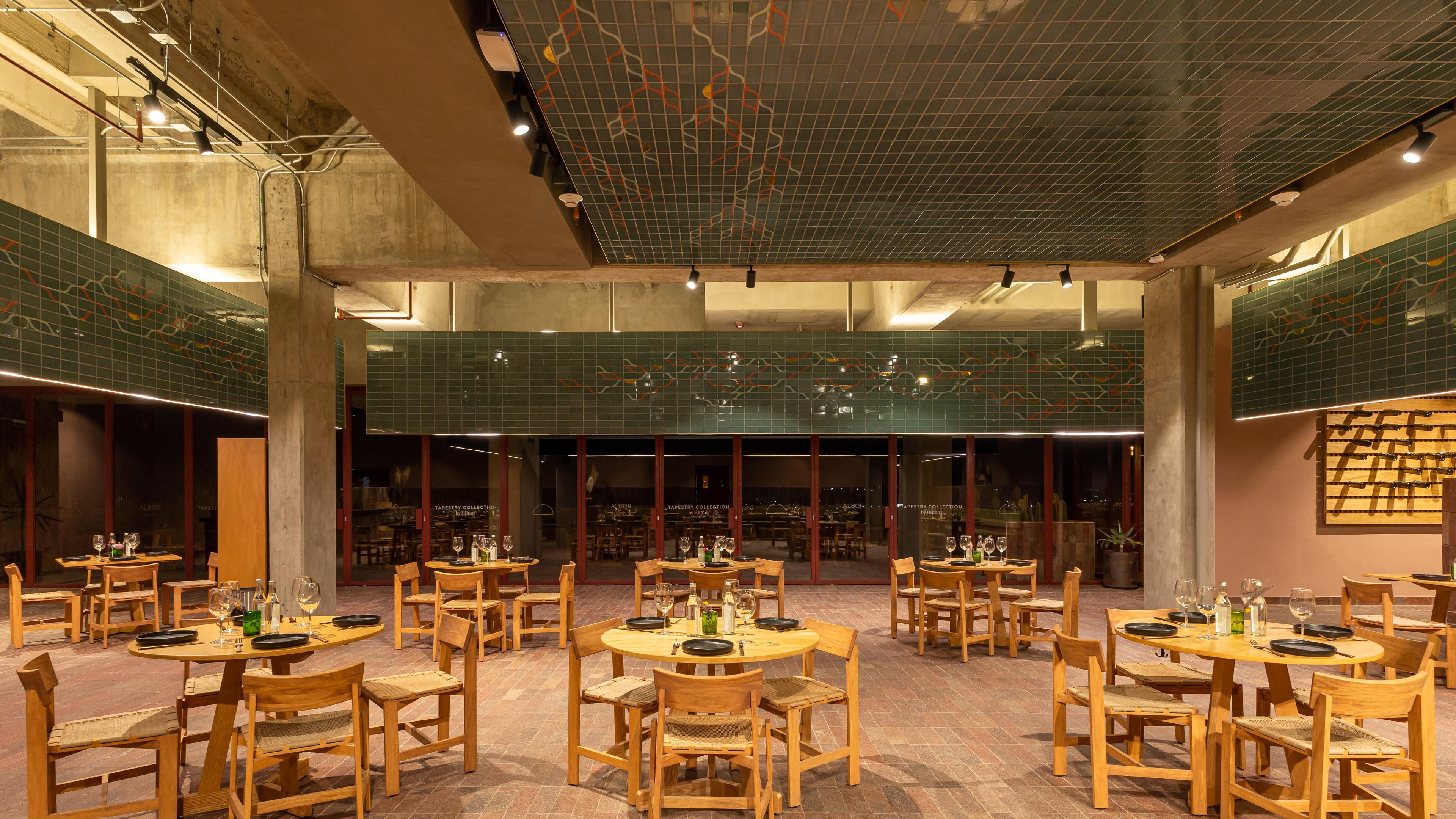 A spacious indoor dining area with multiple round tables and wooden chairs, featuring a high ceiling with a grid pattern and green-tiled walls.