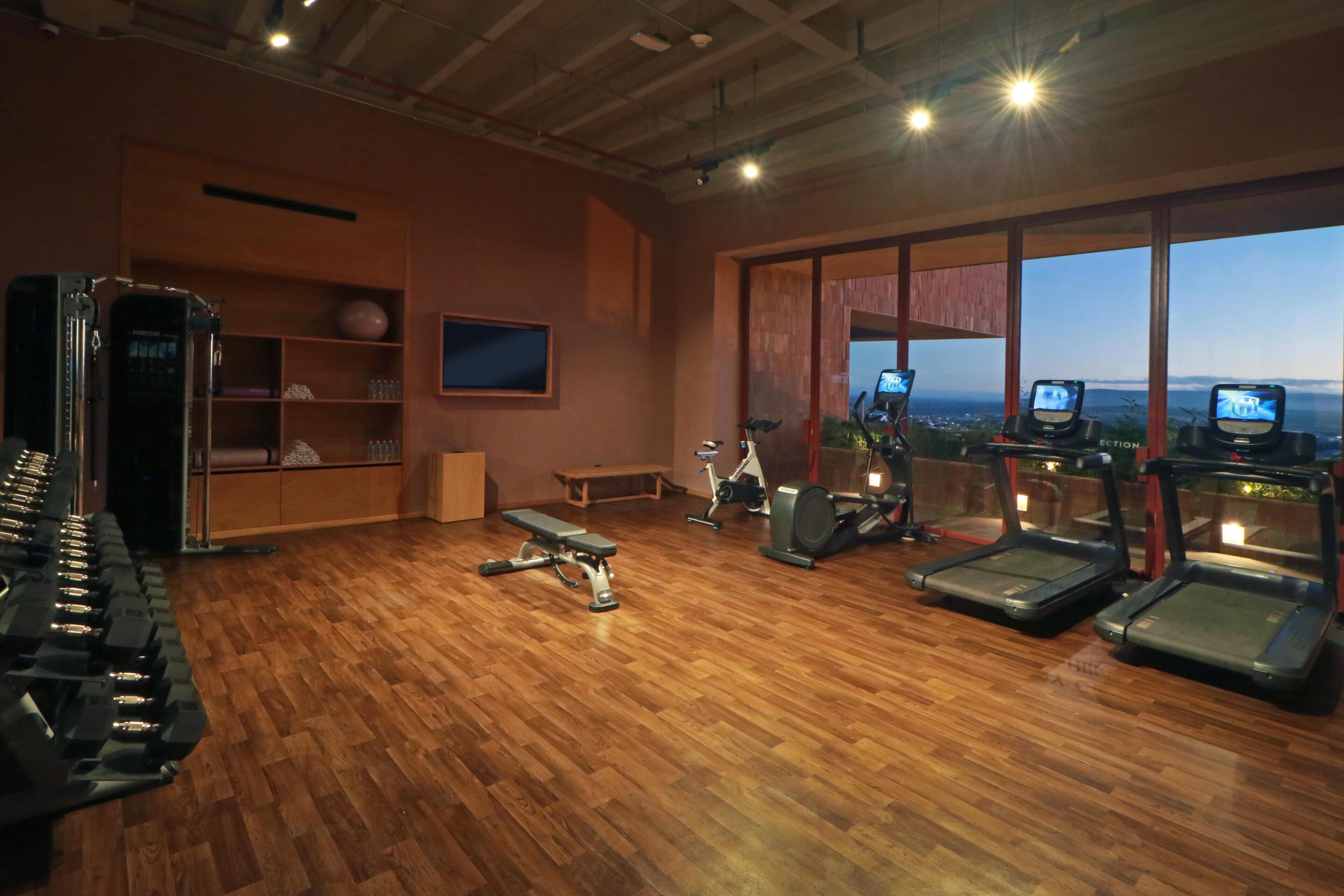 Une salle de sport équipée de tapis de course, d’haltères et d’autres appareils d’exercice. On y trouve également un banc et un téléviseur fixé au mur.