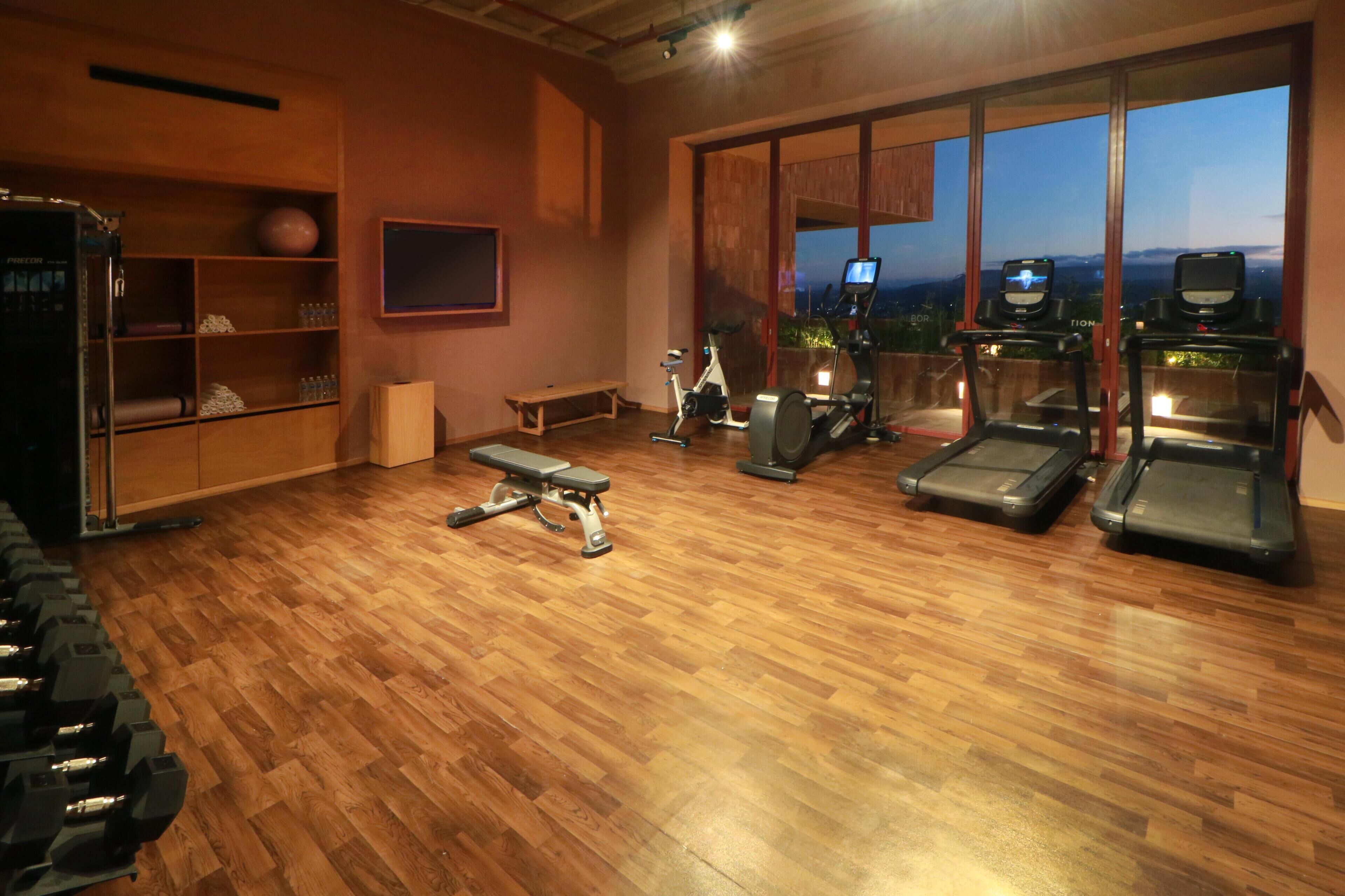 Un gimnasio con equipo de ejercicio, piso de madera y ventanas amplias con vistas.