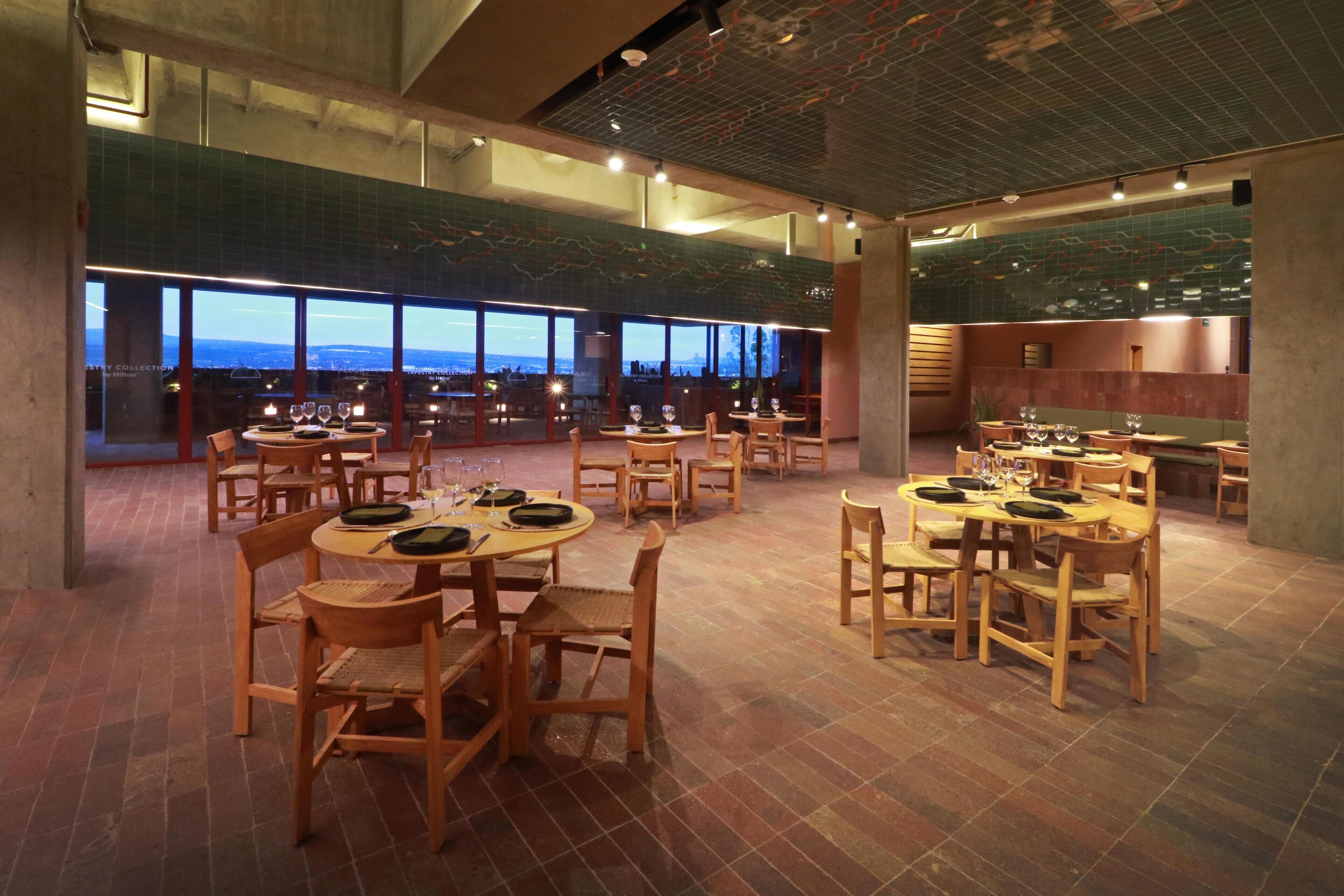 Um restaurante espaçoso com várias mesas e cadeiras, grandes janelas com vista para a paisagem urbana e teto alto com vigas aparentes.