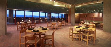 Um restaurante espaçoso com várias mesas e cadeiras, grandes janelas com vista para a paisagem urbana e teto alto com vigas aparentes.