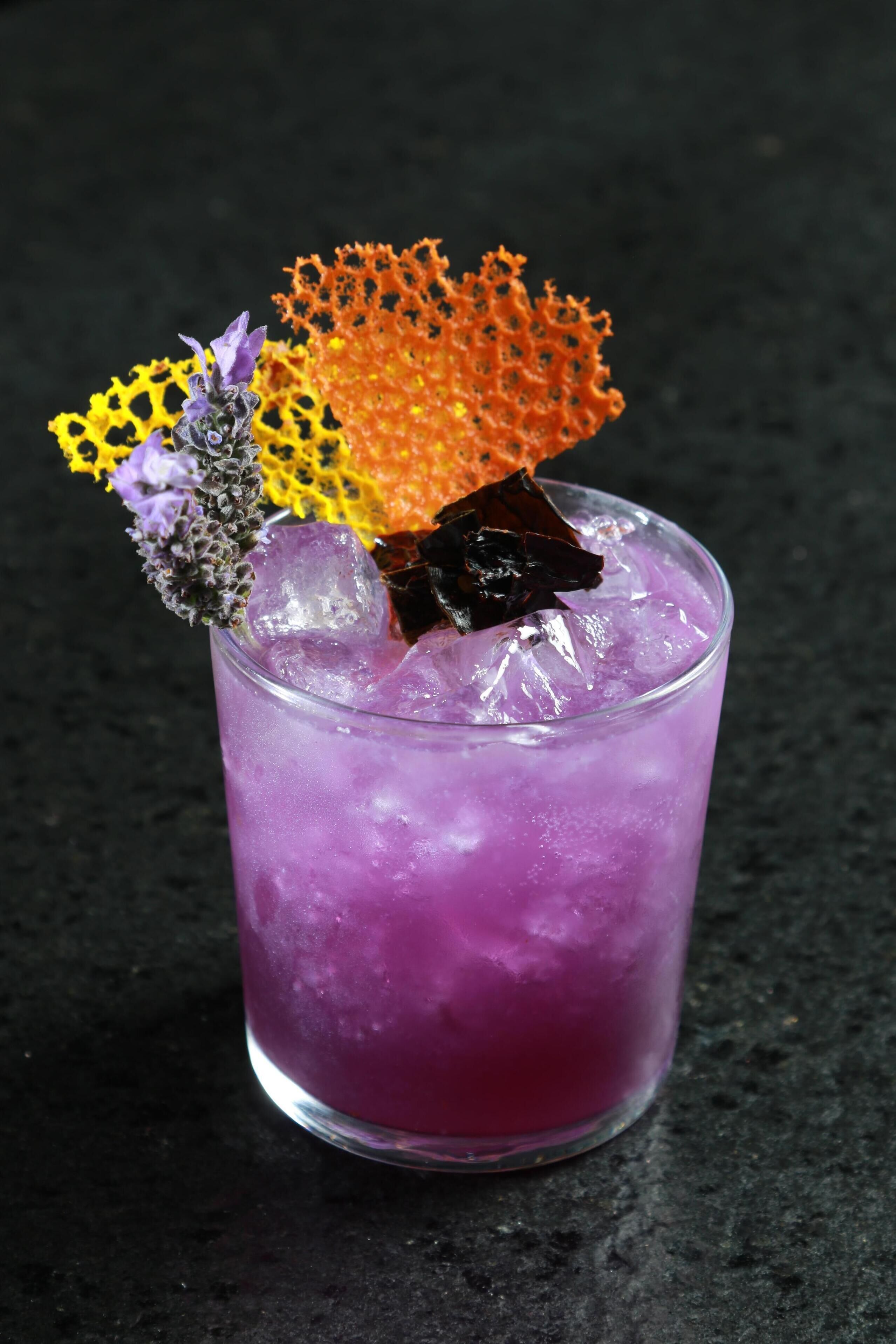 Ein purpurner Cocktail mit Eiswürfeln und Dekoration auf einer dunklen Oberfläche.