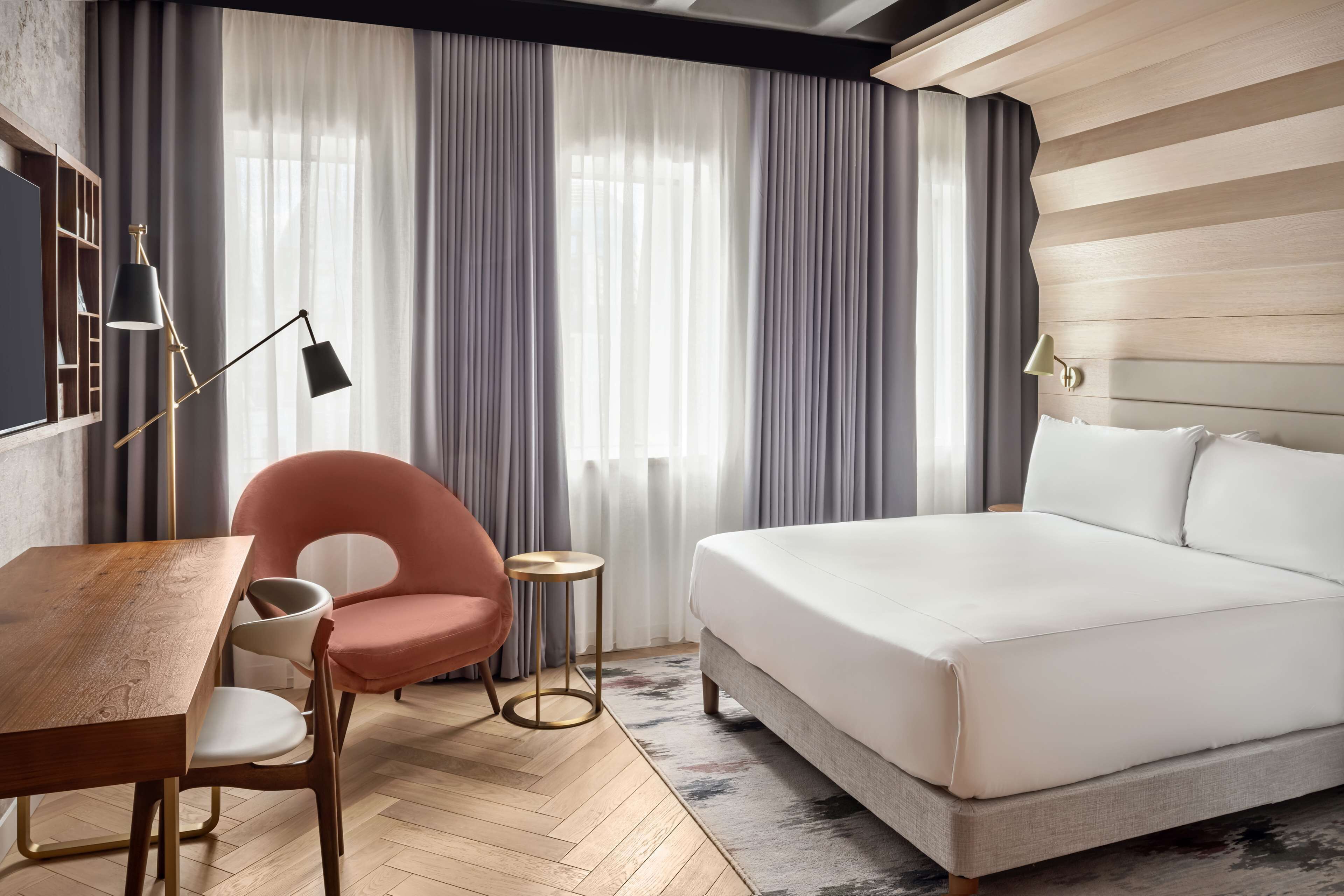 Une chambre d’hôtel moderne avec un plancher en bois, un grand lit, un bureau avec une chaise, une petite table ronde et une chaise rose.