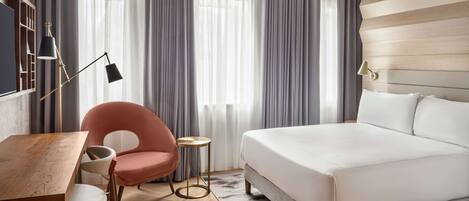 Une chambre d’hôtel moderne avec un plancher en bois, un grand lit, un bureau avec une chaise, une petite table ronde et une chaise rose.