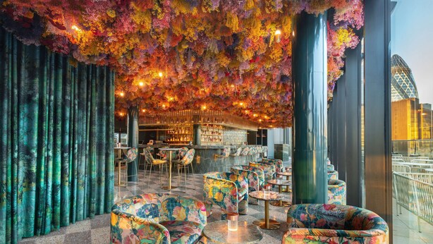 Et moderne barområde med farverige blomsterdekorationer i loftet, behagelig siddeplads og udsigt til en markant bygning i baggrunden.