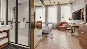 Et moderne hotelværelse med et glasindrammet brusebad, en seng med hvid gavl, et skrivebord med stol og et lille bord med lampe.