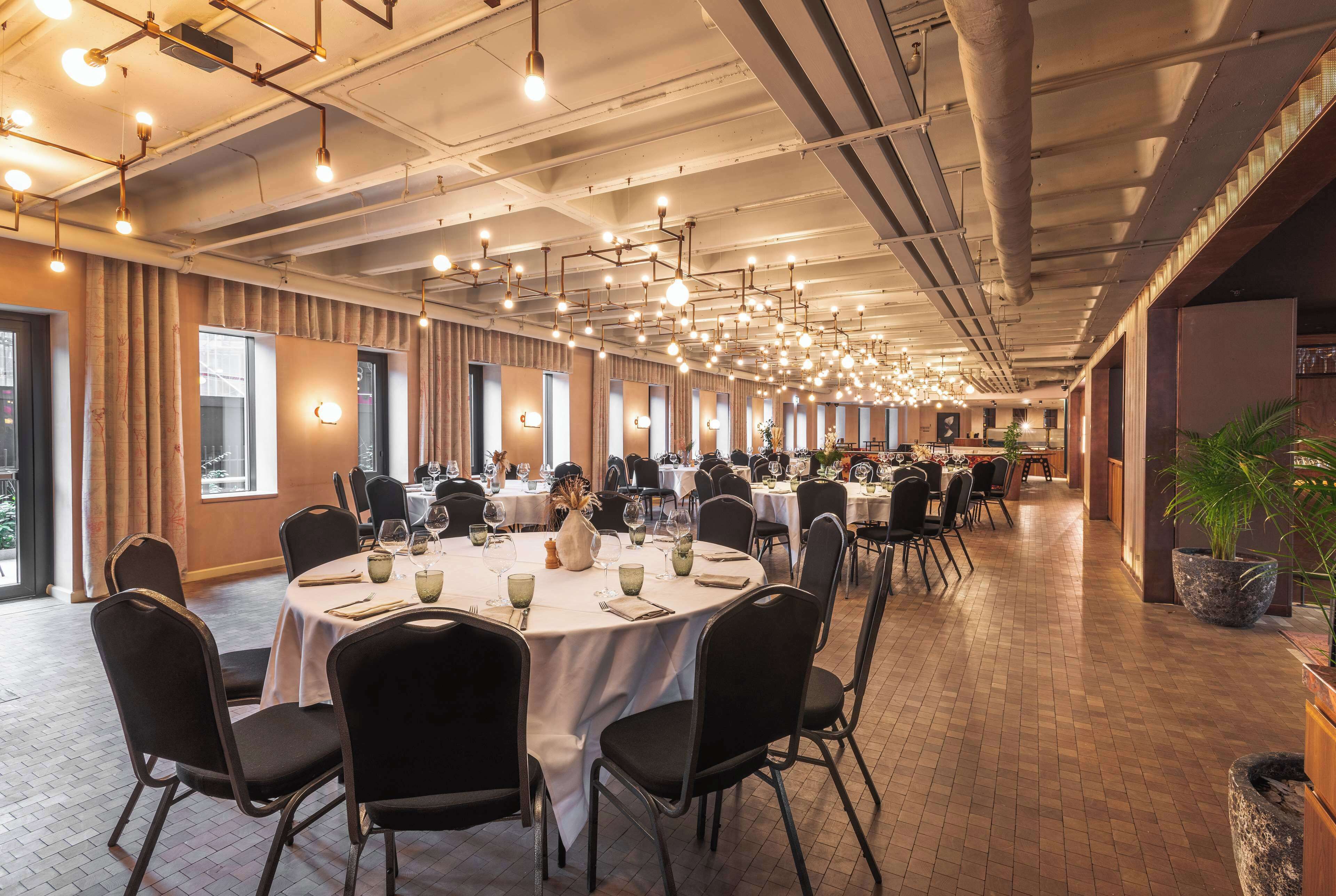 A spacious banquet hall with round tables, black chairs, and hanging pendant lights.