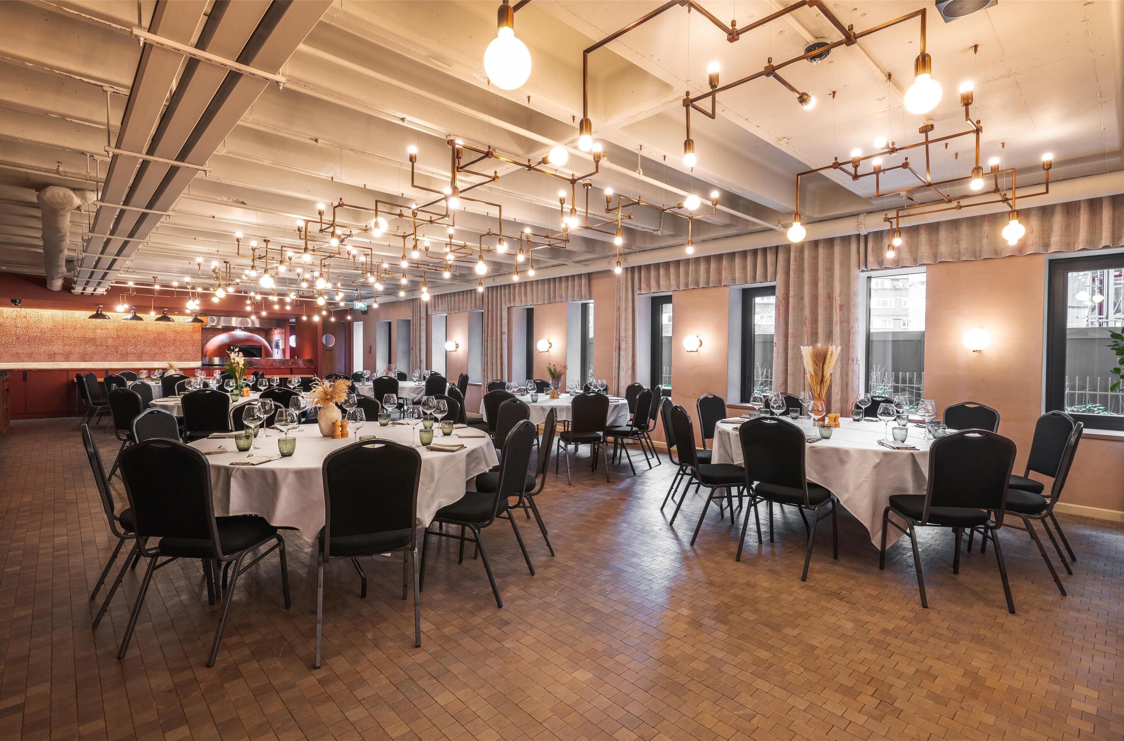 Un'ampia sala da pranzo con diversi tavoli rotondi, sedie nere e un soffitto con tubature a vista e lampade a sospensione.