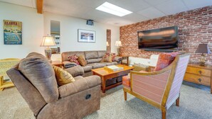 Uma sala de estar com parede de tijolos, uma TV de tela plana, um sofá de couro marrom, uma mesa de centro de madeira e uma poltrona estampada.