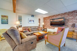 A living room with a brick wall, a flat-screen TV, a brown leather sofa, a wooden coffee table, and a patterned armchair. - Anchorage Apt: Home Base for Alaska Adventure! (Anchorage)