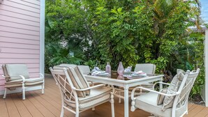 Un patio al aire libre con una mesa y sillas blancas, rodeado de abundante vegetación.