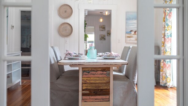 Un comedor con una mesa blanca, sillas tapizadas en gris, una araña y un panel de madera rústica en el centro.