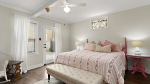 Un dormitorio con una cama grande, un banco, un ventilador de techo, una ventana con cortinas y una mesita de noche con lámpara.