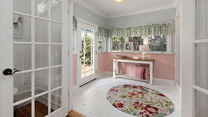 Una habitación con un amplio ventanal, suelo de madera, alfombra redonda con motivos florales, una mesita con una cesta y un banco rosa.