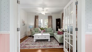 Un salón con ventilador de techo blanco, sofá verde, mesa de centro blanca y cortinas con estampado floral.