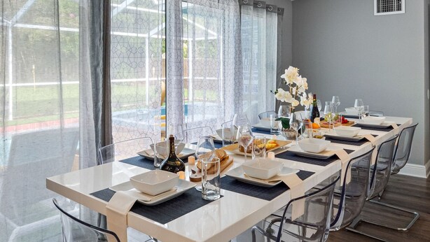 Ein Essbereich mit einem langen Tisch, gedeckten Plätzen für sechs Personen, transparenten Stühlen und Blick auf einen Garten durch große Fenster.