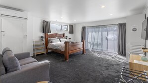 A modern bedroom with a wooden bed, a grey sofa, a desk, and a large window with curtains. - Nine Acres - FREE Wi-Fi (Invercargill)