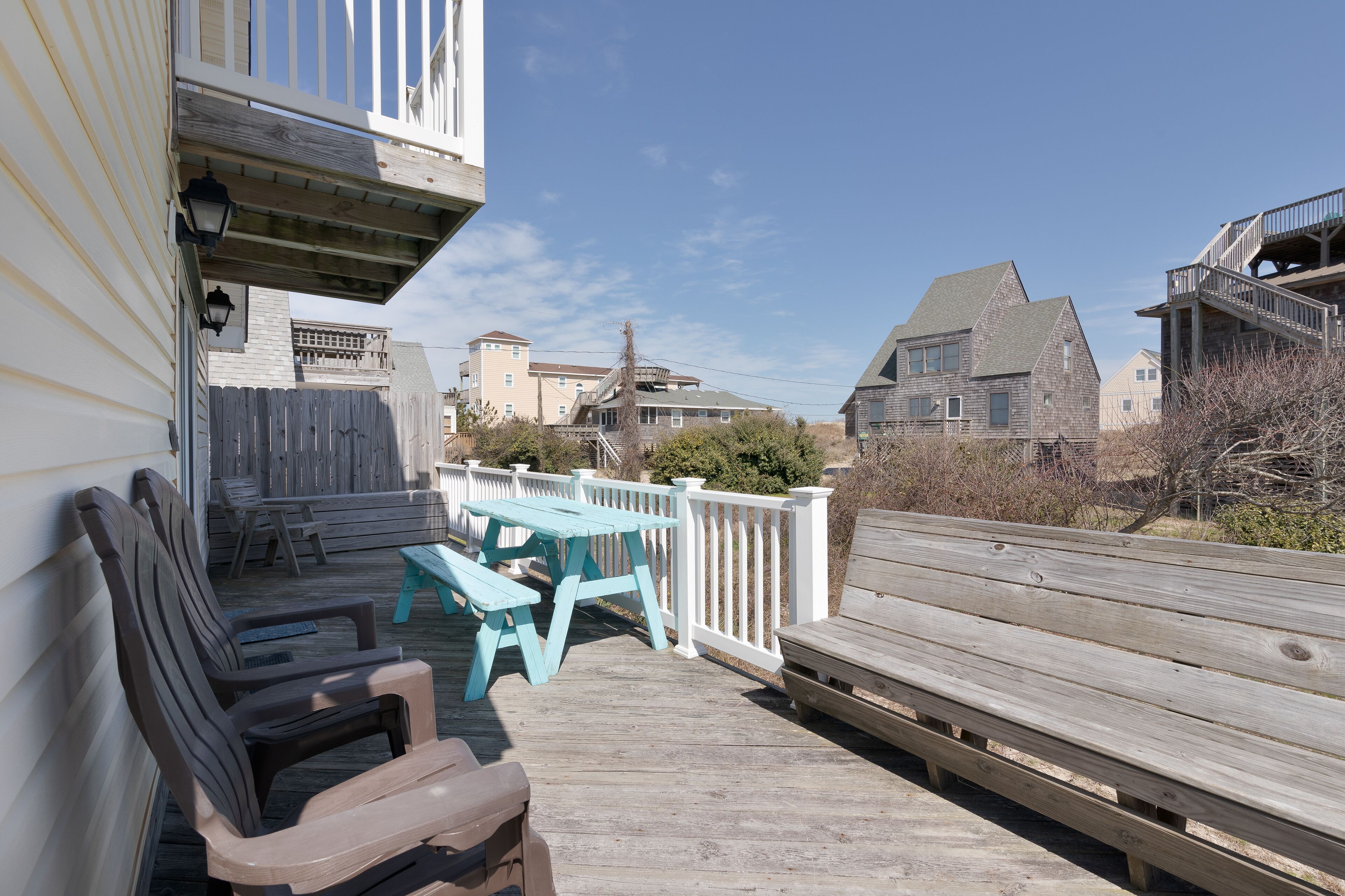 A wooden deck with picnic tables and chairs, a white railing, and a view of surrounding buildings.