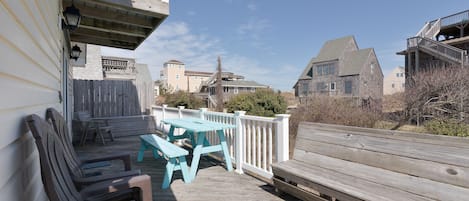 A wooden deck with picnic tables and chairs, a white railing, and a view of surrounding buildings.