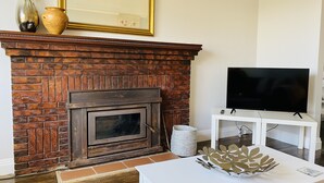 A living room with a brick fireplace, a flat-screen TV on a white stand, and a decorative metal leaf sculpture on a white coffee table. - Daisy Cottage on Duke (Yarram)