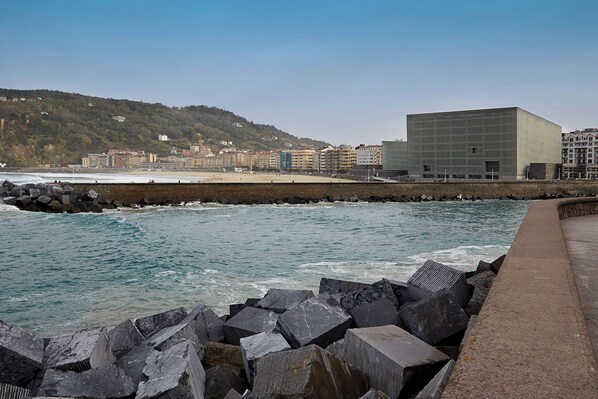 A coastal cityscape with a modern building, rocky breakwaters, and a beach in the background.