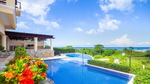 Una piscina con vista al océano, rodeada de vegetación y un edificio con terraza techada.