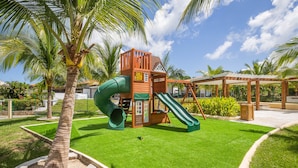 Un parque infantil con tobogán y estructura para escalar, rodeado de palmeras y un césped cuidado.