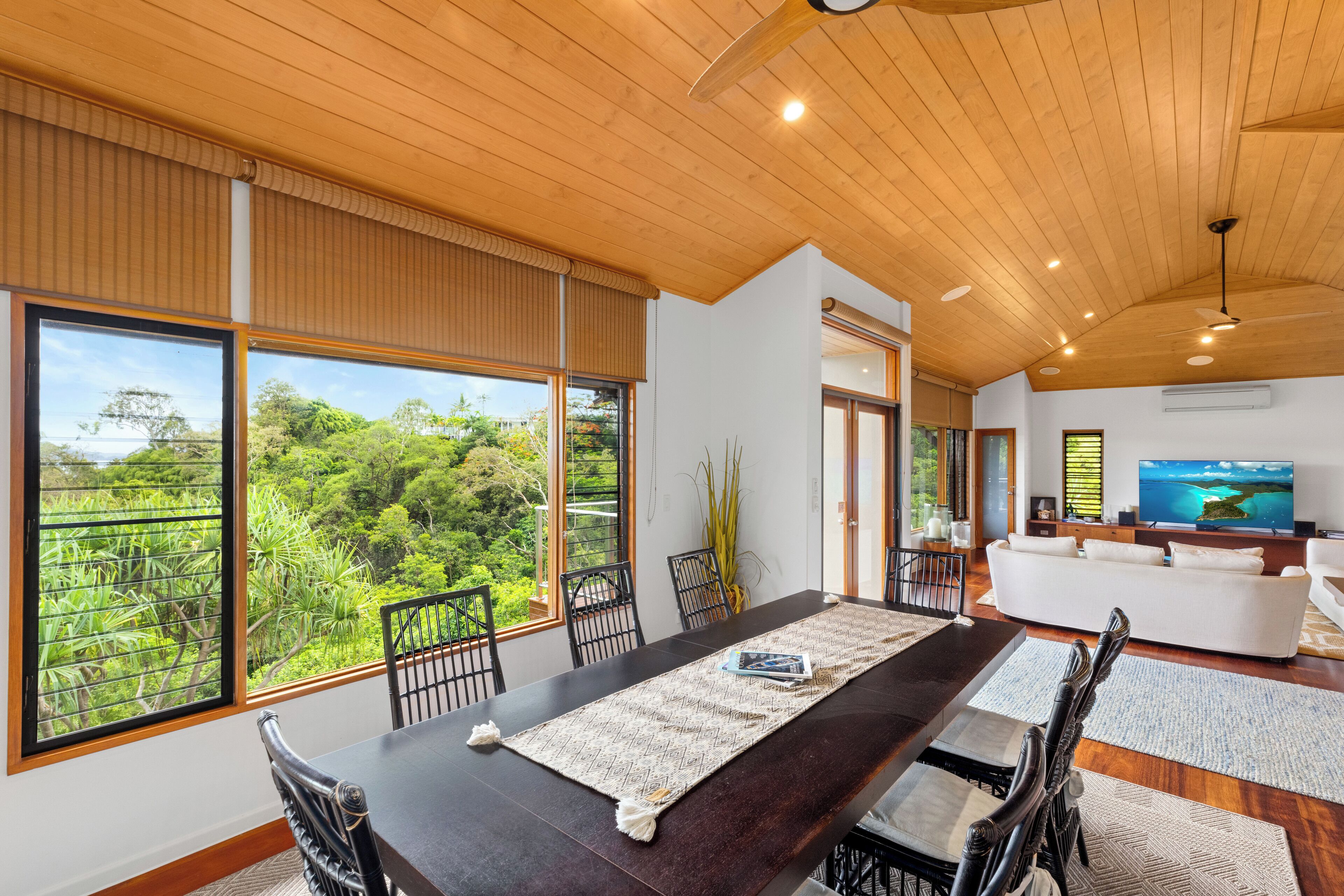Un salon spacieux avec une grande table à manger en bois, des chaises noires et un canapé blanc. Une grande fenêtre offre une vue sur la verdure luxuriante à l’extérieur.