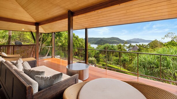 A wooden deck with a sofa and round table, overlooking a lush landscape with mountains and water.