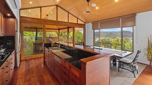 A spacious kitchen with a large island, wooden ceiling, and large windows offering a view of greenery.