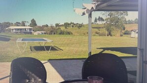 A covered outdoor area with a table and two chairs, a glass of drink, and a view of a grassy field with a building in the distance. - Horotiu Large Group Accommodation & Events (Horotiu)