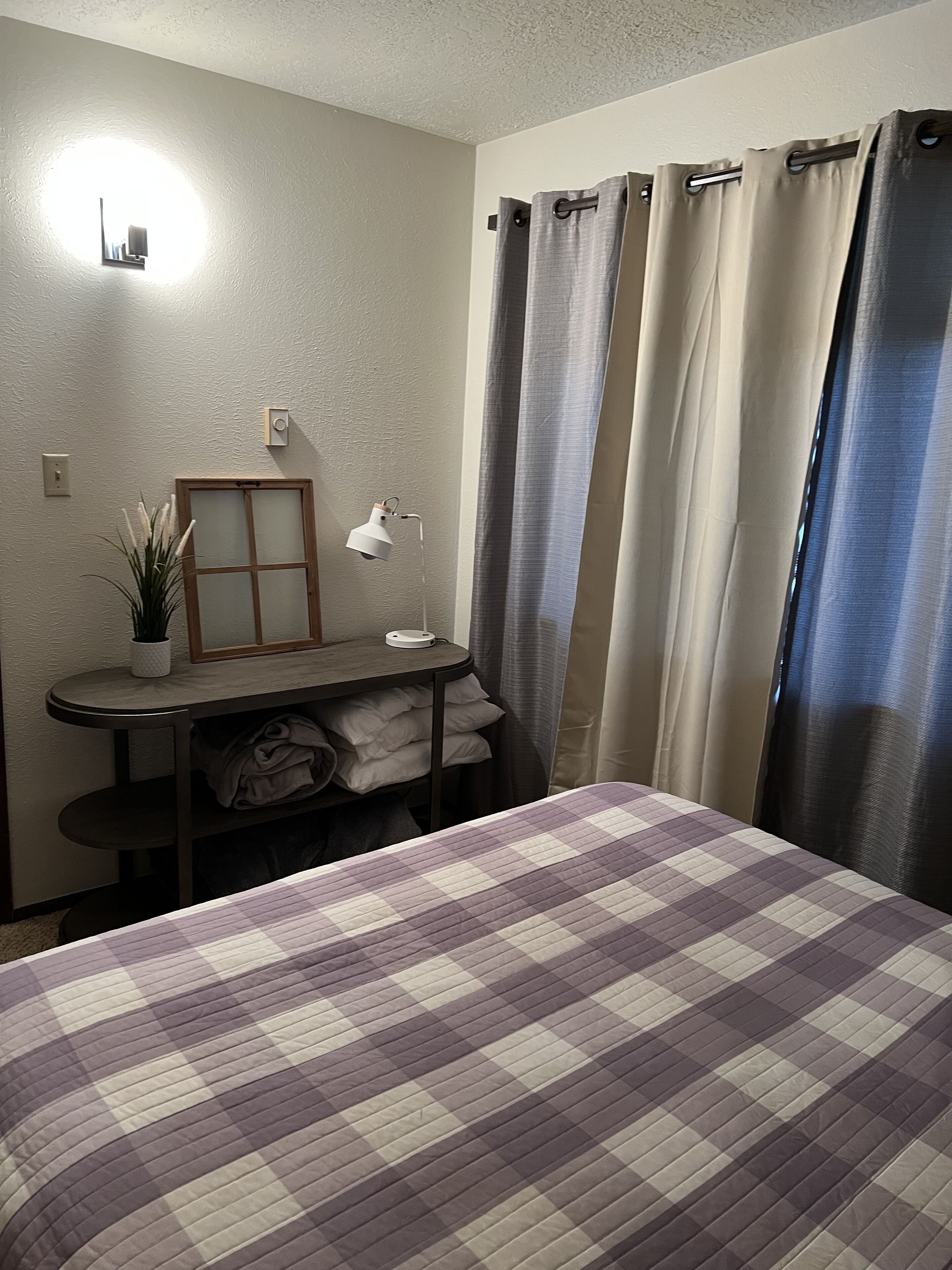 Ein Schlafzimmer mit einem Bett, einem Nachttisch mit Lampe, einer kleinen Topfpflanze und einem Fenster mit Vorhängen.