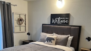 Een slaapkamer met een groot bed, een hoofdeinde, een nachtkastje, een wandlamp, een decoratie aan de muur en een raam met gordijnen.