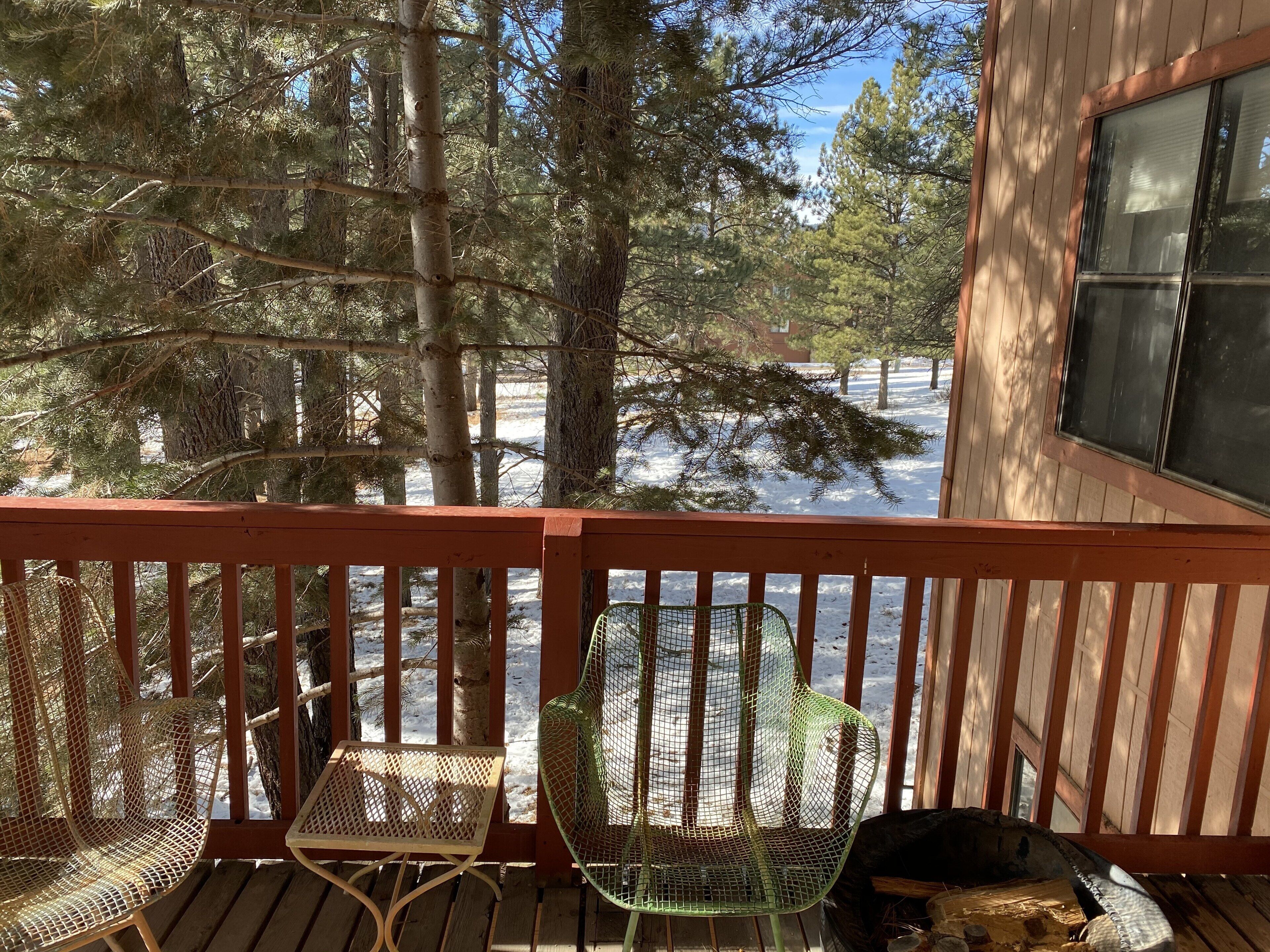 A wooden deck with two wicker chairs and a green mesh chair, overlooking a snowy landscape with trees.