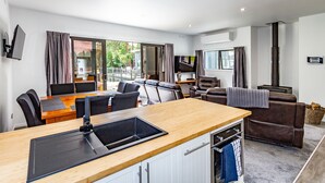 Cozinha moderna com bancada de madeira, pia integrada e armários brancos. Há uma área de jantar com mesa e cadeiras, e um espaço de estar com sofá e poltronas.