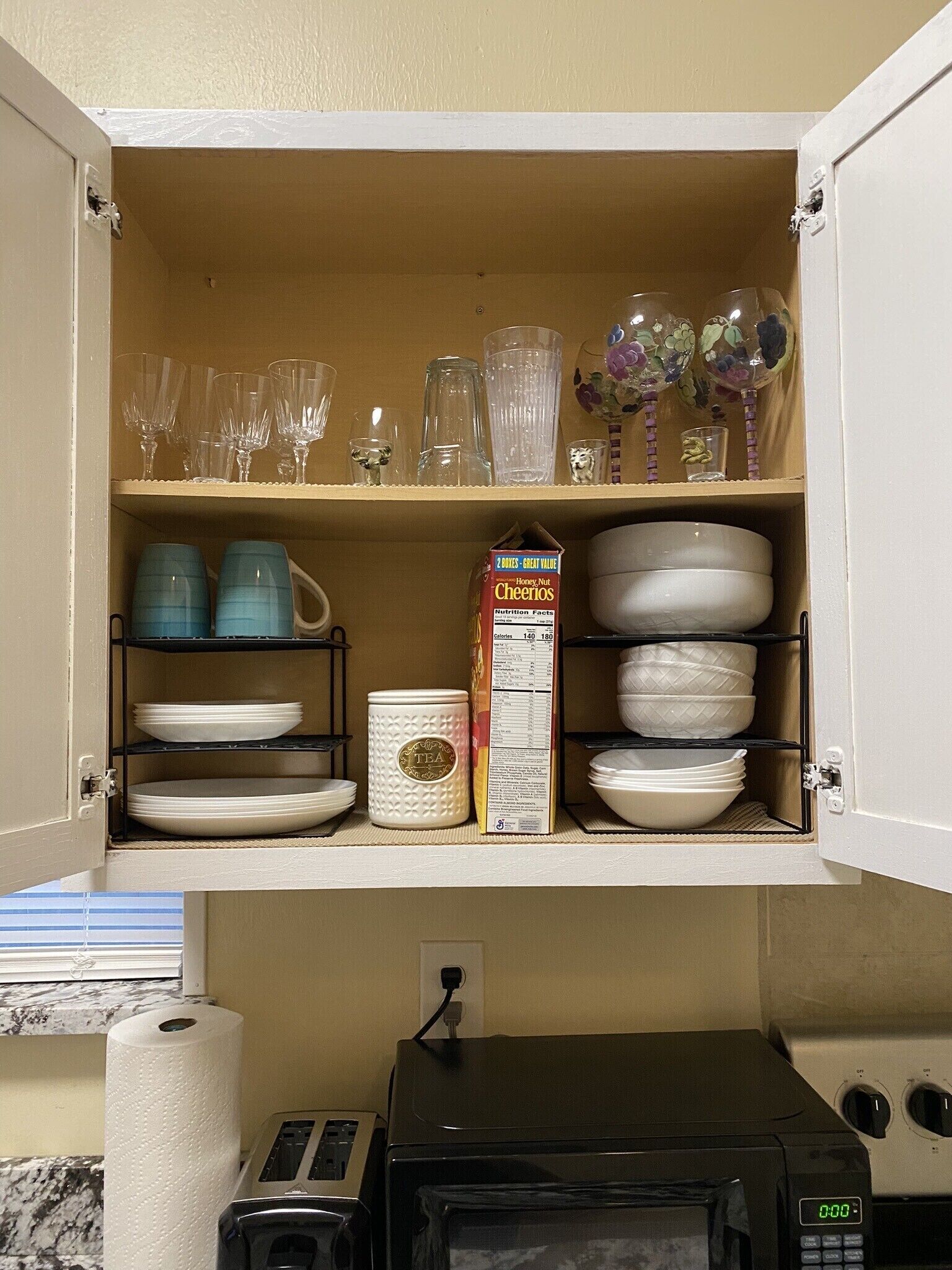 A kitchen cabinet with shelves holding various glassware, plates, and a box of Cheerios cereal.