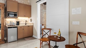Una cocina compacta con microondas, cafetera y lavamanos. Hay una mesa de comedor de madera con dos sillas y una botella de vino. Una puerta corrediza da a otra habitación.